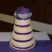 First Wedding Cake Round 4 tier covered in buttercream icing, Purple ribbon accents and sild flowers on top.